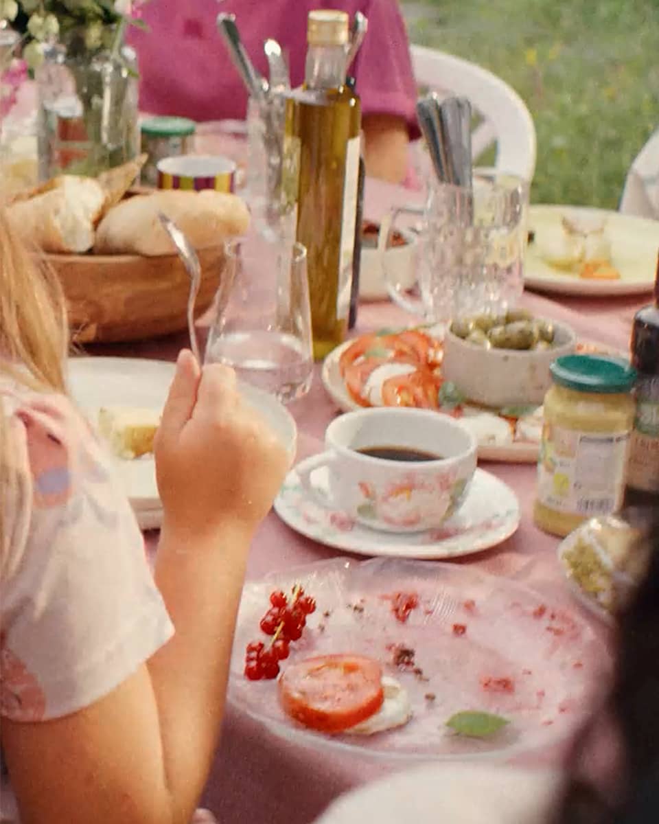 Ein gedeckter Tisch mit einer Tasse Kaffee, einem Teller mit Tomatenscheiben, einem Glas Wasser und einem Korb mit Brötchen. Eine Person hält eine Gabel in der Hand