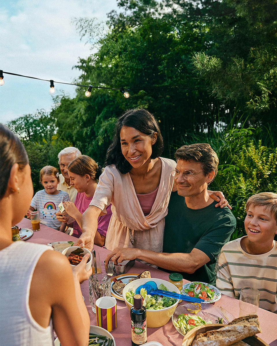 Eine Gruppe von Menschen sitzt an einem Tisch im Freien, auf dem Essen serviert wird, darunter eine Schüssel mit Würstchen. Im Hintergrund sind grüne Pflanzen zu sehen. Lichterkette hängt über dem Tisch.