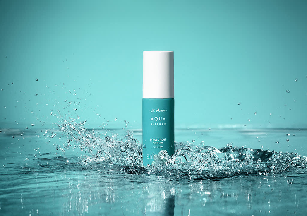 Eine türkisfarbene Flasche mit weißem Deckel und der Aufschrift „Aqua Intense Hyaluron Serum“ steht in spritzendem Wasser