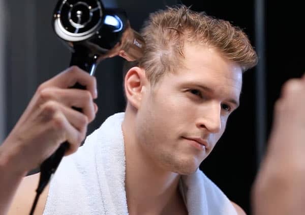 Ein Mann trocknet sein Haar mit einem schwarzen Föhn, ein weißes Handtuch liegt um seine Schultern