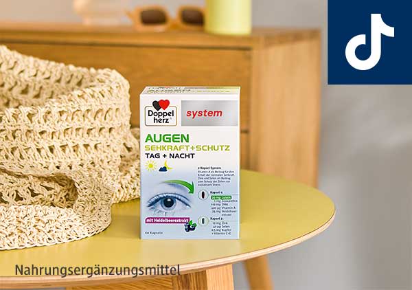 Verpackung des Nahrungsergänzungsmittels Doppelherz System Augen Sehkraft + Schutz Tag + Nacht auf einem gelben Tisch, daneben ein gestricktes Tuch. Unten im Bild steht „Nahrungsergänzungsmittel“