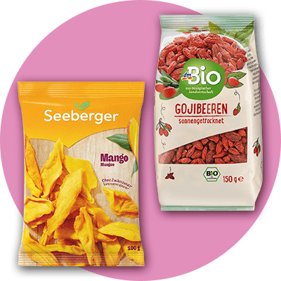 Eine gelbe Verpackung von Seeberger mit getrockneten Mangostücken und der Aufschrift „Mango“ neben einer weißen dmBio-Packung mit getrockneten Gojibeeren und der Aufschrift „Gojibeeren sonnengetrocknet“ auf einem rosa Hintergrund