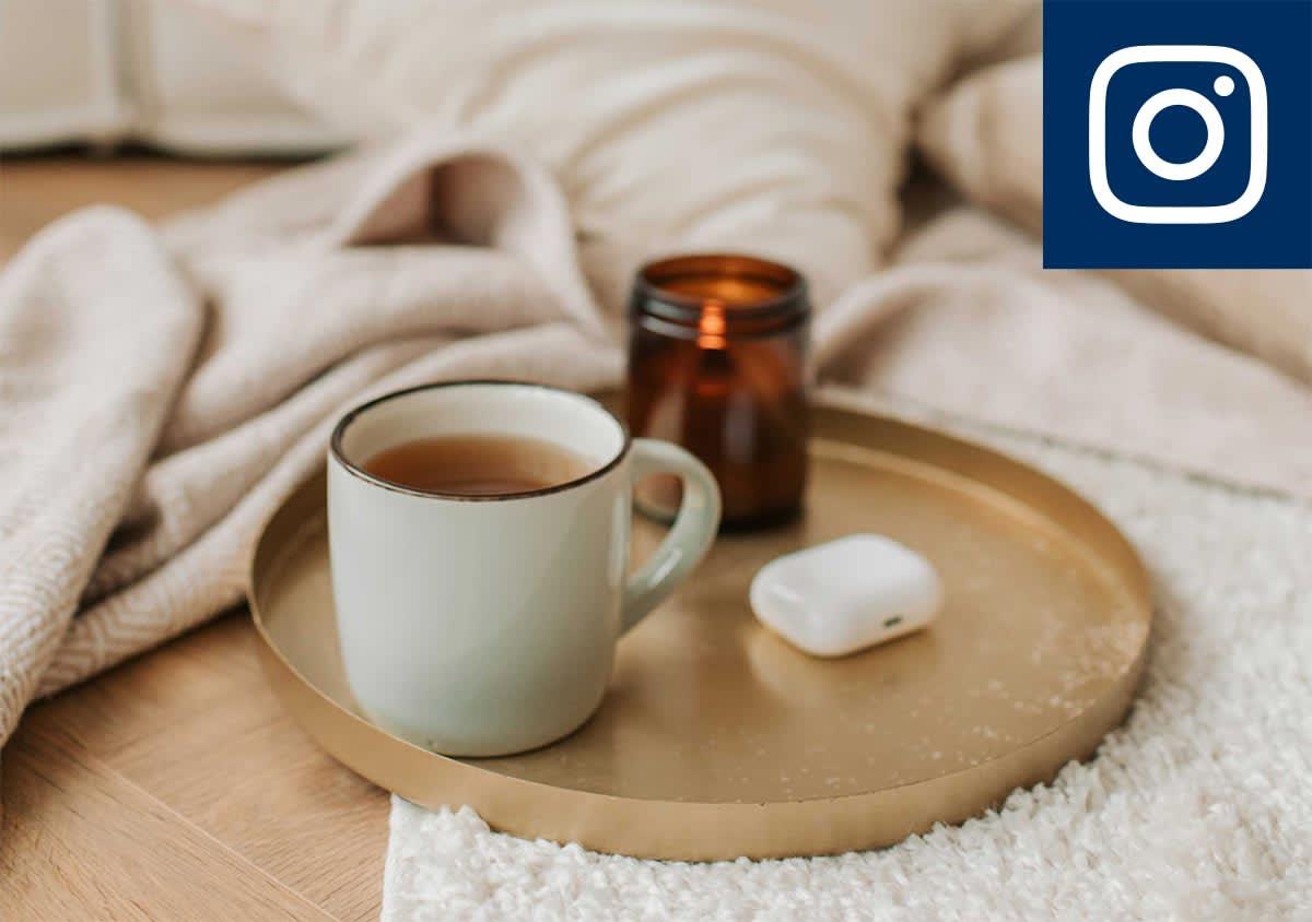 Eine Tasse Tee, ein braunes Glas und ein weißes kabelloses Kopfhörer-Etui auf einem goldenen Tablett, umgeben von weichen Decken auf einem Holzboden