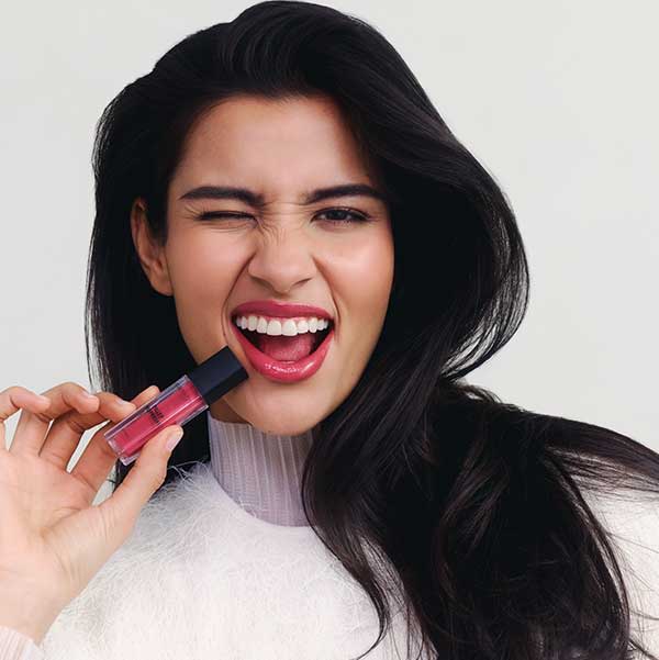 Eine Person hält einen Lippenstift in der Hand, der eine kräftige rosa Farbe hat. Die Person trägt ein weißes Oberteil mit einem hohen Kragen.
