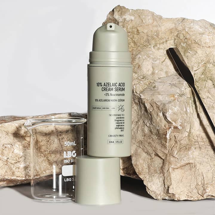 Eine graue Flasche mit der Aufschrift „10% Azelaic Acid Cream Serum“ steht vor einem steinigen Hintergrund. Daneben ein Becherglas mit der Aufschrift „50ml LBG ISO 3“
