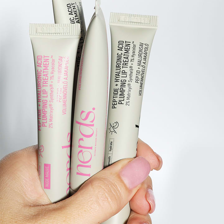 Drei Tuben des Produkts „Peptide + Hyaluronic Acid Plumping Lip Treatment“ von nerds werden in einer Hand gehalten. Die Tuben sind weiß mit pinker und schwarzer Schrift.