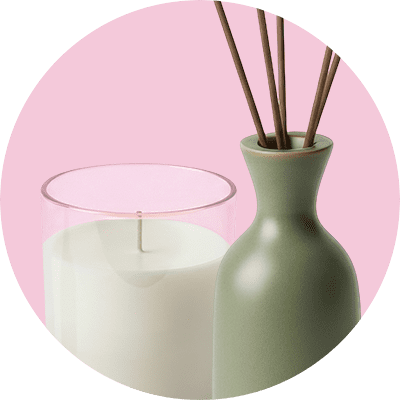 Eine brennende weiße Kerze in einem durchsichtigen Glas neben einer mattschlanken, grünen Duftspender-Vase mit drei dünnen Rührstäbchen in einem pastellrosa Kreishintergrund