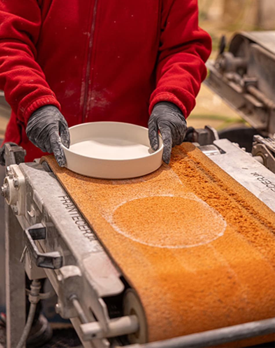 Eine Person in rotem Pullover und schwarzen Handschuhen hält eine runde weiße Schale über eine auf einer Maschine laufende Schicht aus feinem orangebraunem Sand