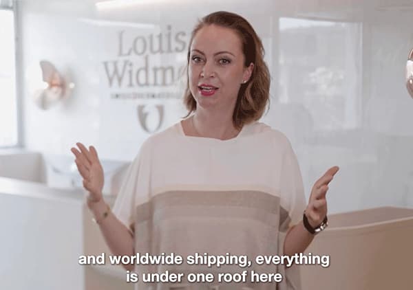 Frau steht vor einer Glaswand mit dem Schriftzug „Louise Widmer“, spricht und gestikuliert, eingeblendeter englischer Untertitel „and worldwide shipping, everything is under one roof here“