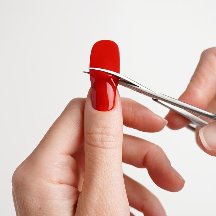 HĂ€nde halten Daumen mit roten PressâOnâNagel; mit einer Nagelschere wird ĂŒberschĂŒssiger NagelrĂŒcken gekĂŒrzt