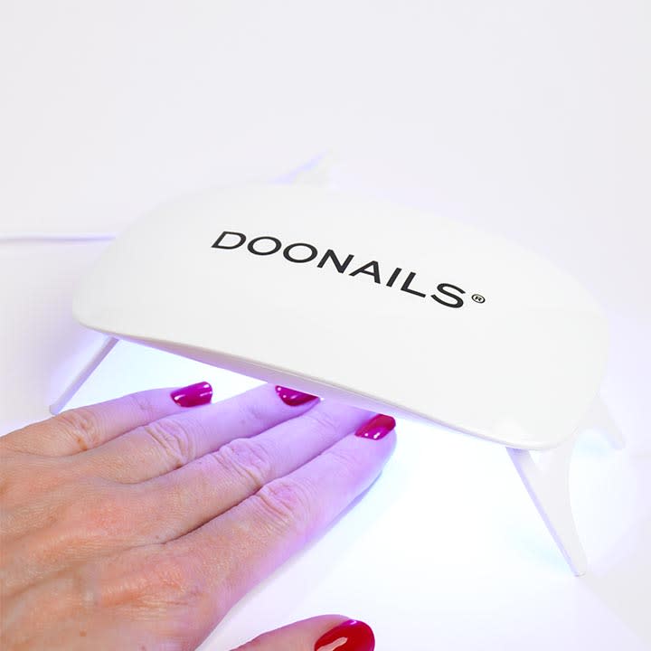 Hand mit lackierten roten FingernĂ€geln steckt unter einer weiĂen UV-/LED-Nageltrockner-Lampe mit schwarzem Schriftzug âDOONAILSâ