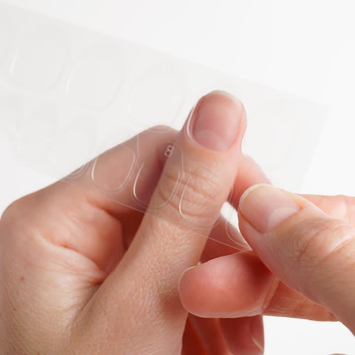 HÀnde halten eine transparente Folie mit selbstklebenden, ovalen NagelaufsÀtzen und lösen einen Aufsatz ab