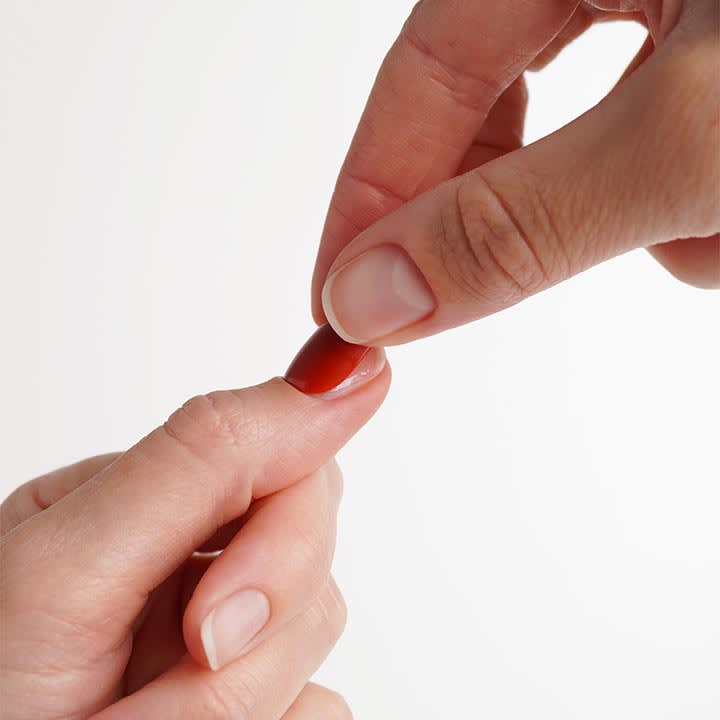 Zwei HÀnde, eine Hand hÀlt einen roten Weichkapsel-VitaminprÀparat zwischen Daumen und Zeigefinger und setzt es auf den ausgestreckten Zeigefinger der anderen Hand an