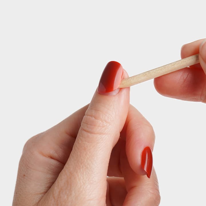 HÀnde mit roten, glÀnzenden lackierten NÀgeln, eine Hand verwendet ein HolzstÀbchen, um Nagellack am Daumennagel zu formen oder zu sÀubern