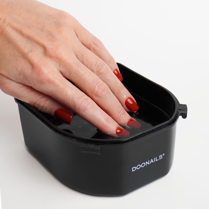 Hand mit roten lackierten FingernĂ€geln taucht Finger in schwarzem NagelbadbehĂ€lter mit Aufschrift âDOONAILSâ