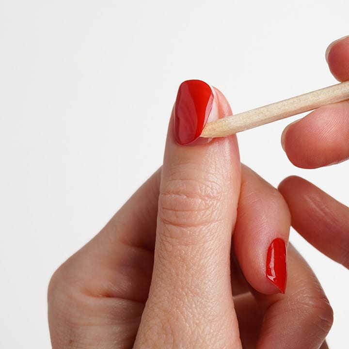 HÀnde mit rot lackierten FingernÀgeln, mit einem HolzstÀbchen wird Nagellack am Nagelrand entfernt