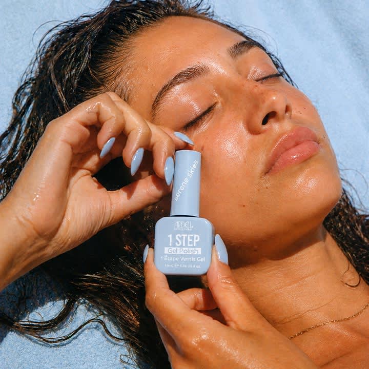 Frau liegend auf blauem Handtuch, mit nassen dunklen Haaren, hält nahe Hals und Kinn eine kleine hellblaue Nagellack-Verpackung mit Aufschrift „1 STEP Gel Polish“, sichtbare lange pastellblaue lackierte Fingernägel
