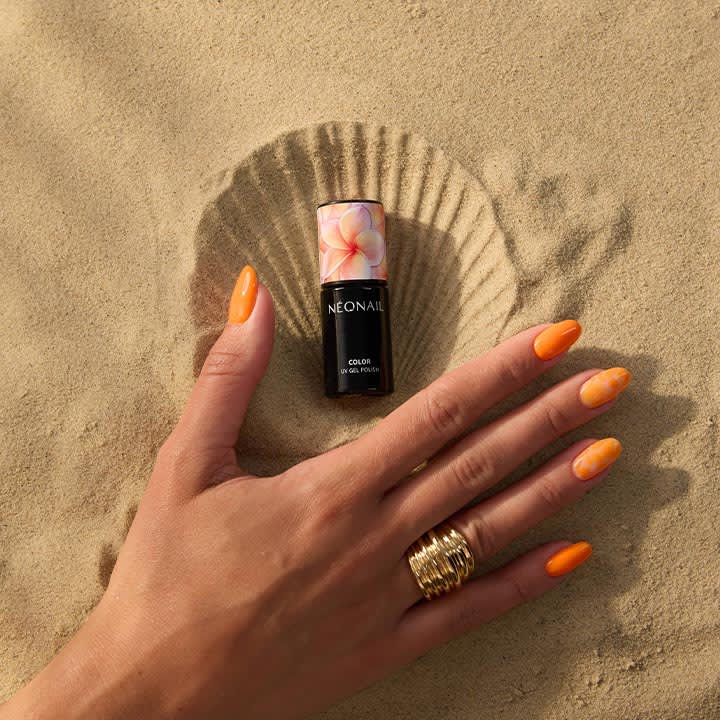 rechte Hand mit orangen, mandelförmigen Gelnägeln und mehreren goldenen Ringen liegt in Sand, darüber eine schwarz-weiße Nagellack-Flasche mit Blumendesign und Schriftzug NEONAIL