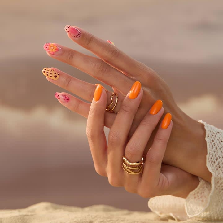 Zwei überkreuzte Hände mit langen, orangen und gemusterten rosa Gelnägeln, mehrere goldene Ringe an den Fingern, helles Häkelärmelsaum sichtbar