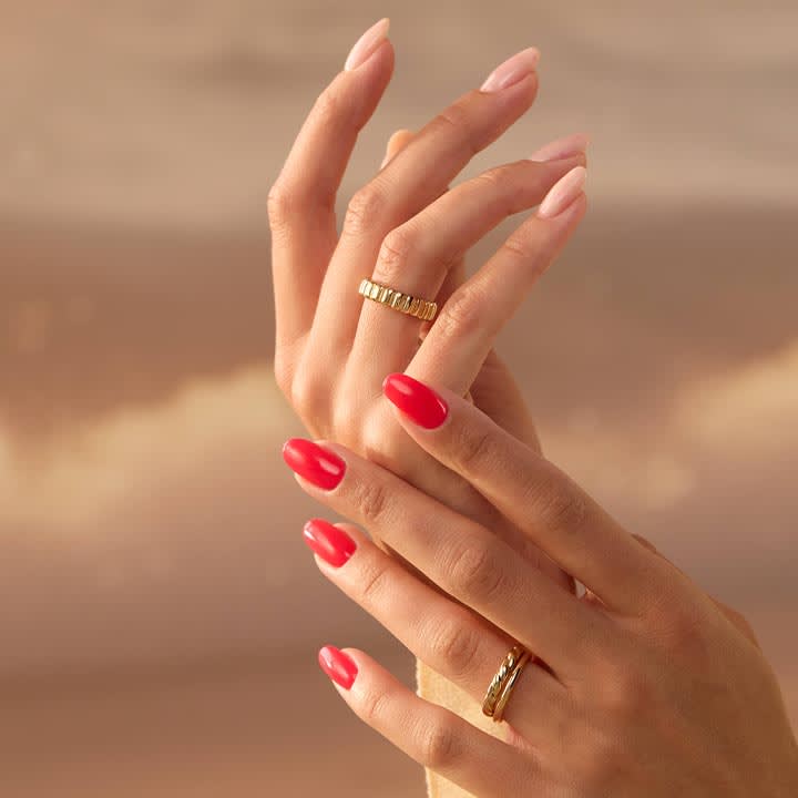 Zwei übereinandergelegte Hände mit roten und nude lackierten Nägeln, jeweils ein goldener Ring an Mittelfinger und Ringfinger sichtbar