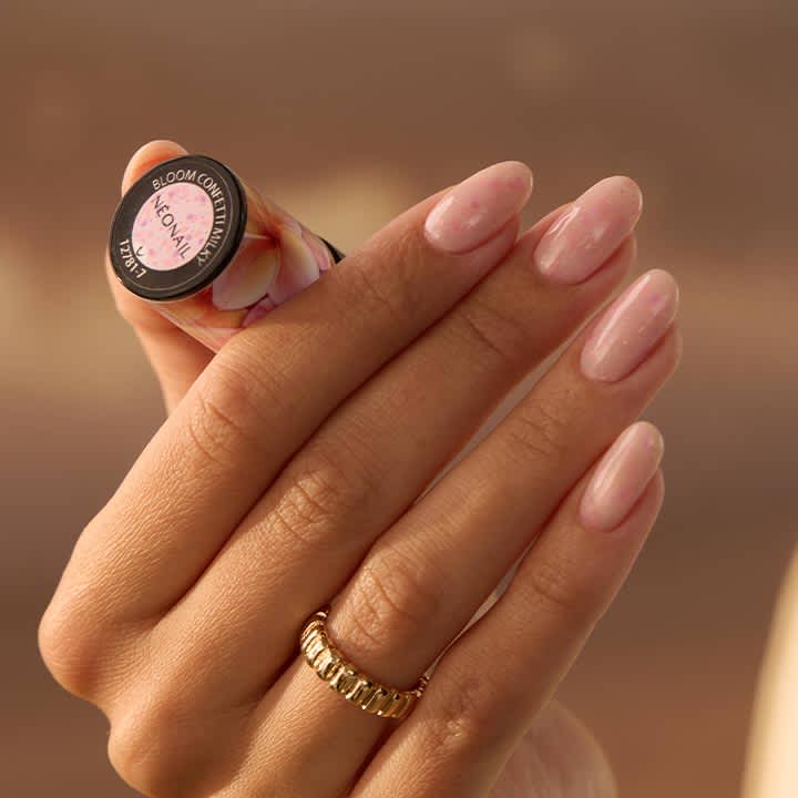 Hand mit langen, ovalen, pastellrosa lackierten Fingernägeln hält eine kleine Nagellack-Flasche; goldener Ring am Ringfinger sichtbar
