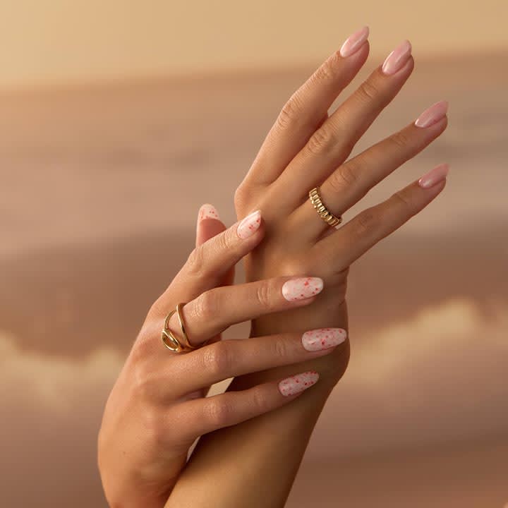 Zwei überkreuzte Hände mit gepflegten, länglichen Nägeln; die oberen Nägel sind rosa lackiert, die unteren rosa mit weißen Sprenkeln, an den Fingern goldene Ringe