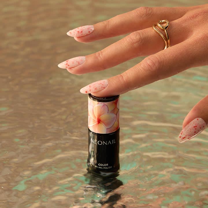 Hand mit längeren, weiß-rosa gesprenkelten Gelnägeln und goldfarbenem Ring berührt eine aufrechter stehende schwarze Nagellackflasche mit bunter Blütenetikette und Marke „NEONAIL“