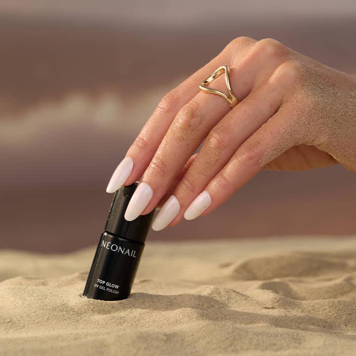 Hand mit sandbedeckten Fingern und langen, hellrosa lackierten Nägeln hält eine schwarze Nagellackflasche mit Aufschrift „NEONAIL“ und darunter „TOP GLOW UV GEL POLISH“; goldener Dreieck-Ring am Mittelfinger