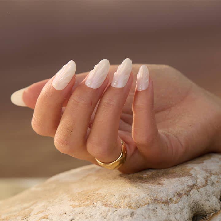 Hand mit hellrosa, oval gefeilten Gelnägeln, drei Nägel mit dezentem Perlmuttmuster, goldener Ring am Ringfinger, Hand liegt auf hellem Stein