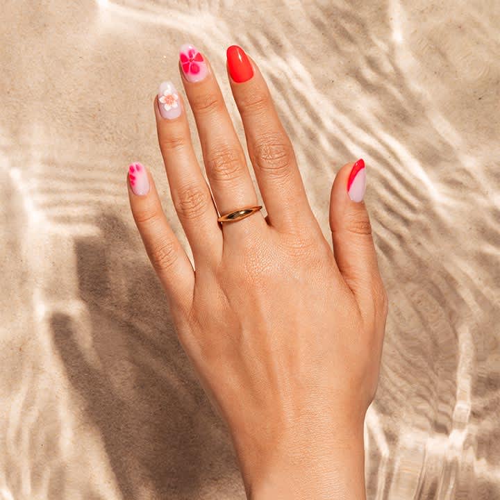 rechte Hand mit goldfarbenem Ring auf dem Mittelfinger, Fingernägel in Pink- und Rottönen lackiert, zwei Nägel mit Blumen-Design, helle Sand- und Wasseroberfläche im Hintergrund