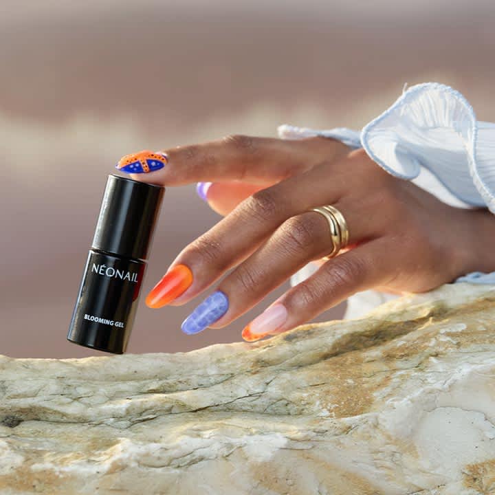 Hand mit farbig gestalteten Fingernägeln (Orange, Lila mit Marmorierung, Rosa) hält eine schwarze Nagelprodukt-Flasche mit der Aufschrift „NEONAIL BLOOMING GEL“ auf einem hellen Steinrand