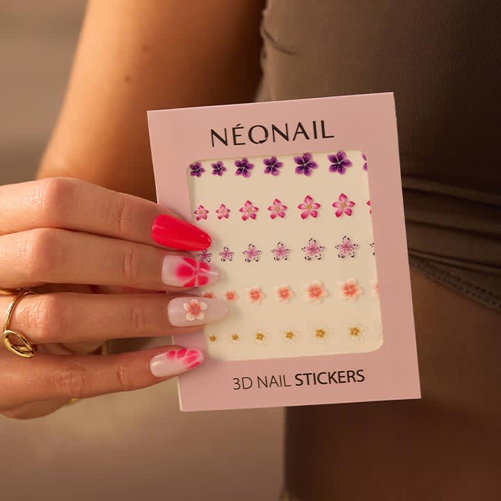 Hand, die eine rosa Verpackung mit der Aufschrift „NÉONAIL“ und „3D NAIL STICKERS“ hält; durchsichtiges Fenster zeigt Reihen kleiner 3D-Nagelsticker in Form violetter, pinker, oranger und goldener Blüten; Hand mit kunstvoll gestylten langen Nägeln in Rot, Weiß und mit Blumenmotiven, trägt goldenen Ring