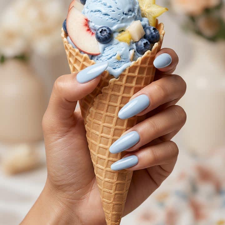 Hand mit hellblauen, langen Fingernägeln hält Waffelhörnchen mit hellblauem Eis, garniert mit Blaubeeren und einer Pfirsichscheibe