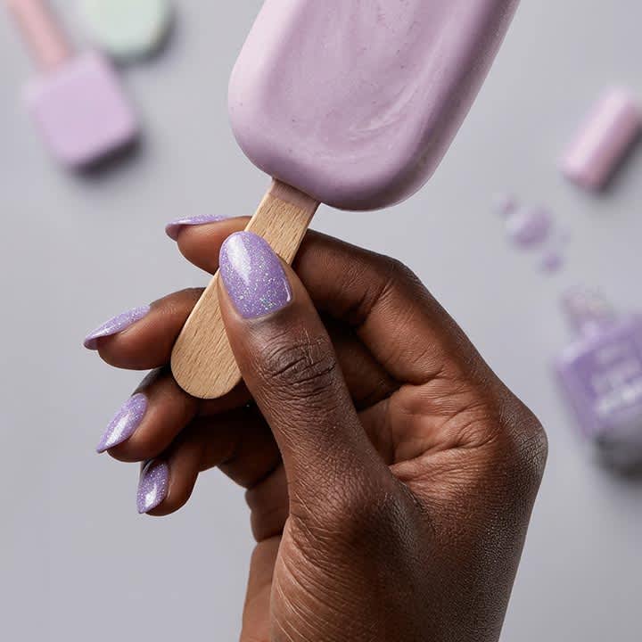 Hand mit dunkler Hautfarbe hält ein lavendelfarbenes Eis am Stiel, Fingernägel lackiert in glitzerndem Flieder, im unscharfen Hintergrund mehrere Nagellackfläschchen und Nagelzubehör in Pastellfarben