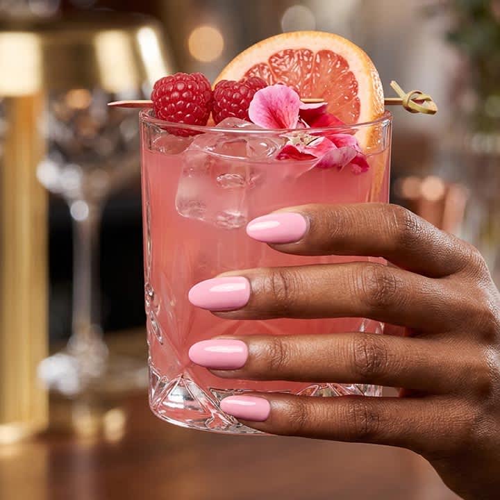 Hand mit rosa lackierten Fingernägeln hält ein Glas mit rosa Cocktail, Eiswürfeln, einer Grapefruitscheibe, Himbeeren und einer kleinen Blüte als Garnitur