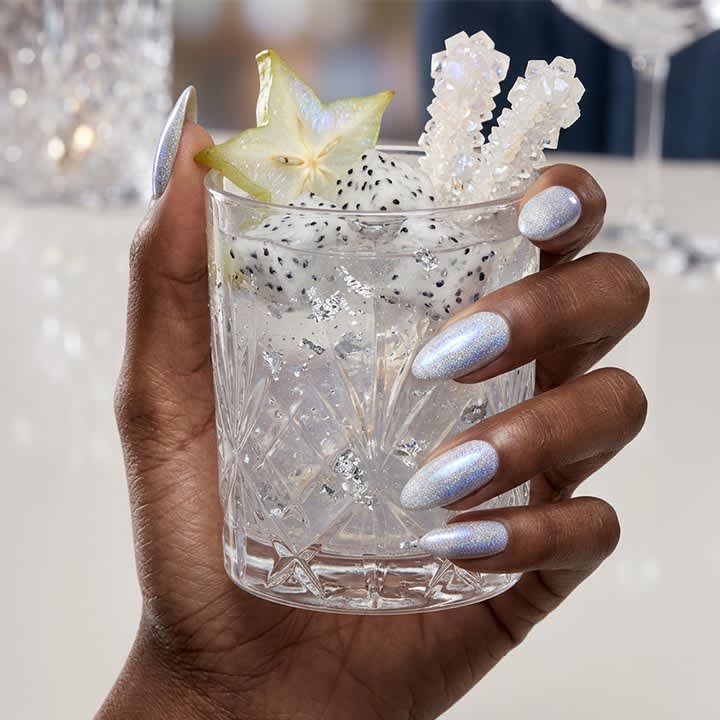 Dunkle Hand mit silbermetallic lackierten Fingernägeln hält ein Kristallglas mit klarem Getränk, Eiswürfeln, zwei weißen Kandiszucker-Sticks und einer halbierten Karambolafrucht am Glasrand