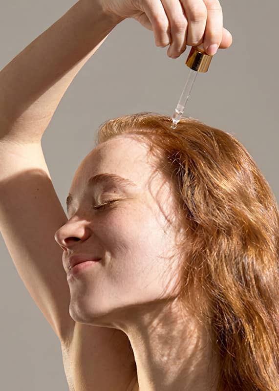 Person füllt mit einer Glaspipette und Goldkappe einen Tropfen Pflegeöl auf den oberen Teil ihrer roten, welligen Haare, Arm erhoben