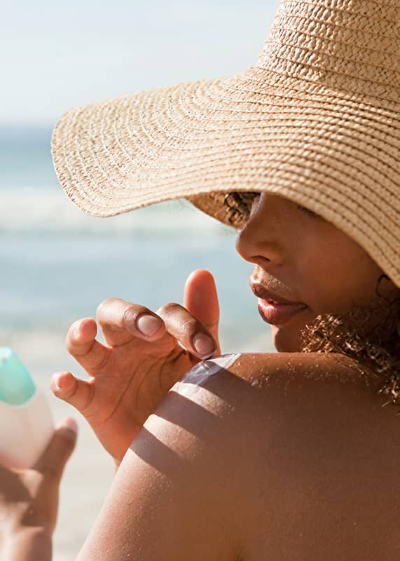 Frau mit Strohhut am Strand, sie hält eine weiße Sonnenschutz-Flasche und trägt Sonnencreme auf ihre Schulter auf