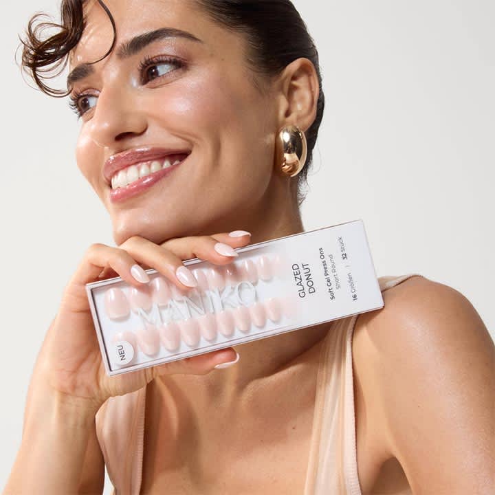Frau hält lächelnd eine transparente Kunststoffverpackung mit künstlichen, hellrosa Fingernägeln und der Aufschrift „GLAZED DONUT“ nahe am Kinn, sichtbare Ohrringe und ärmelloses Top