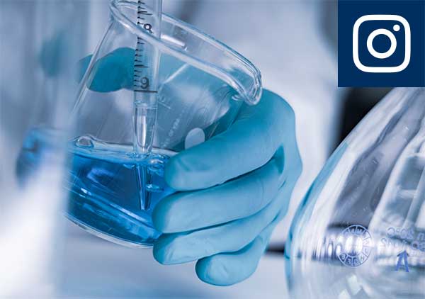 Laborhandschuh behandschuhte Hand hält ein Glasbecherglas mit blauem Flüssigkeitsansatz, daneben ein rundkolben und oben rechts ein Instagram-Logo