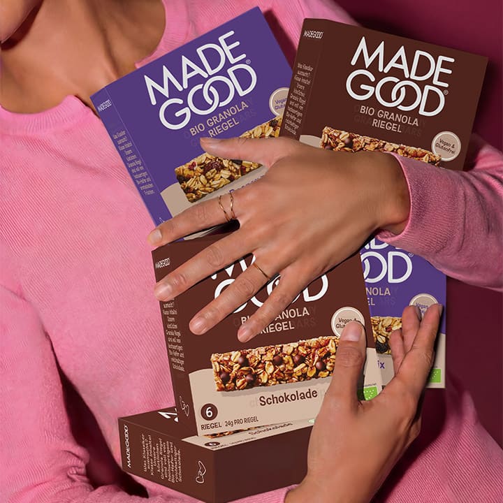 Person in rosa Pullover hält mehrere Packungen Made Good Bio-Granola-Riegel, sichtbare Packungsaufschrift Made Good, Bio Granola Riegel, Geschmacksangabe Schokolade und Packungsfarbe braun und lila, Hände mit Fingerringen um die Packungen