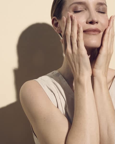 Oberkörper einer Frau in ärmelloser Bluse, die mit beiden Händen Gesicht und Kinn berührt, Blick leicht nach oben gerichtet