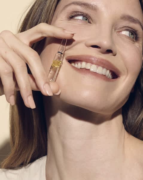 Frau lächelt, hält eine kleine Glasampulle mit gelbem Serum dicht an die Wange