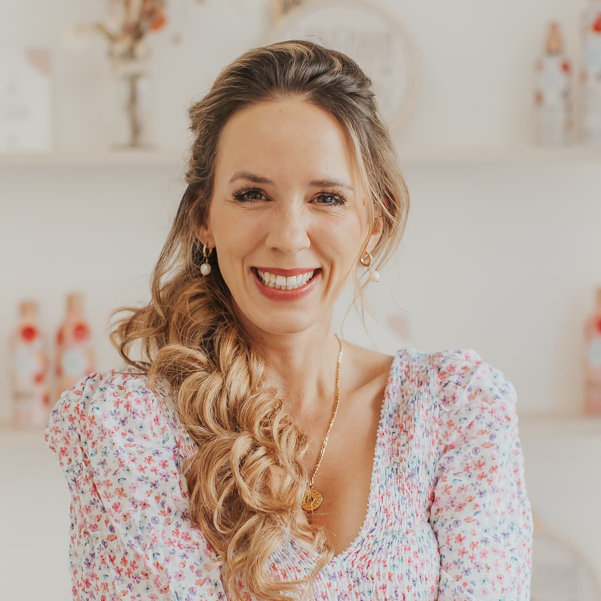 Porträt einer lächelnden Frau mit seitlich geflochtenem langem Haar, floralem Kleid, goldener Kette und Perlenohrringen, vor hellem Regal mit Deko und Produktflaschen
