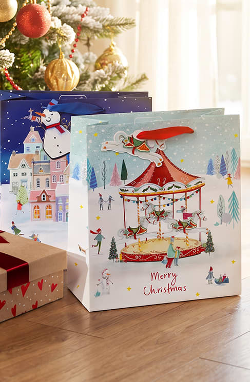 Zwei weihnachtliche Geschenktüten mit Illustrationen; vorne eine hellblaue Tasche mit Karussell, kleinen Figuren und Schriftzug „Merry Christmas“, dahinter eine dunkelblaue Tasche mit Schneemann vor Häusern, links unten ein Geschenkpaket mit rotem Band. Bild mithilfe von KI erstellt