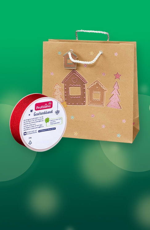 Braunes Geschenkpapier-Tragetasche mit aufgedruckten Lebkuchenhäusern und Tannenbäumen daneben rote Geschenkband-Rolle mit weißem Etikett und Aufschrift „Geschenkband“