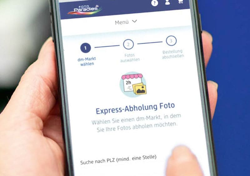 Eine Hand hält ein Smartphone, auf dessen Bildschirm die Webseite von Foto-Paradies geöffnet ist, oben sind das Logo und das Wort Menü zu sehen, darunter sind drei Schritte dargestellt: 1 dm-Markt wählen, 2 Fotos auswählen und 3 Bestellung abschließen, darunter steht Express-Abholung Foto mit einem Symbol eines Ladens und der Aufschrift 2h, darunter der Text Wählen Sie einen dm-Markt, in dem Sie Ihre Fotos abholen möchten, am unteren Rand steht Suche nach PLZ (mind. eine Stelle)