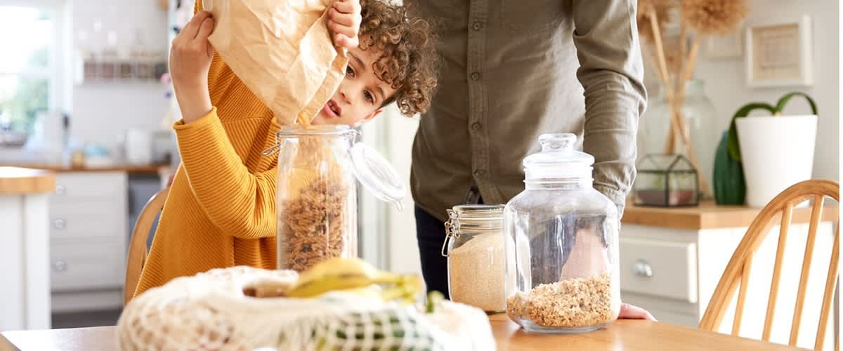 Kind hält Papiertüte über Glasbehälter und schüttet Müsli in ein Vorratsglas, daneben weitere Glasbehälter mit Körnern und Obstnetz auf Holztisch