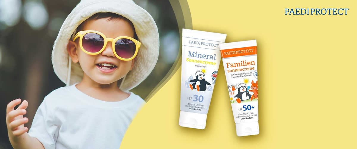Kind lächelnd mit Sonnenhut und gelben Sonnenbrillen, rechts zwei PaediProtect Sonnencreme-Tuben: blaue Tube „Mineral Sonnencreme LSF 30“ mit Pinguin-Motiv und orange Tube „Familien Sonnencreme LSF 50+“ mit Tiermotiven, oben Schriftzug PAEDIPROTECT