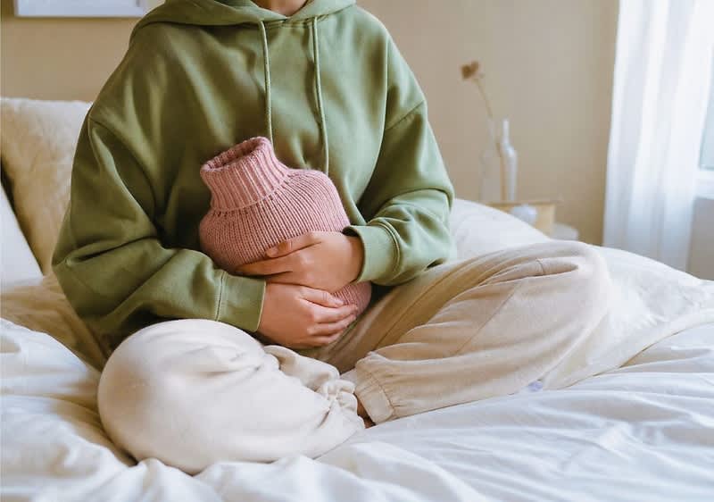Person sitzt im Schneidersitz auf einem Bett und hält eine rosa Wärmflasche mit beiden Händen vor dem Bauch, trägt grünen Hoodie und helle Jogginghose